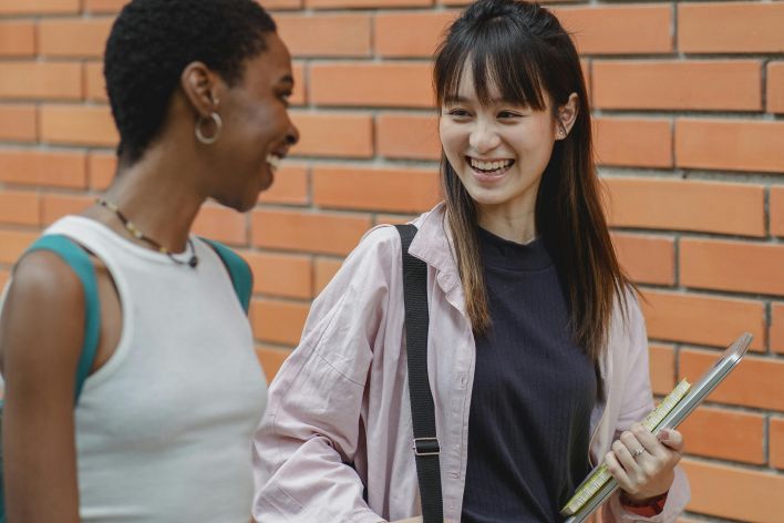 Laughing multiethnic female students wearing casual outfits strolling together against brick wall and chatting happily while looking at each other with smiles
